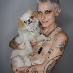 Frankie is standing facing the camera holding their small, fluffy dog, lemon. Frankie’s hair is short and bleached. They are wearing a tank top and black leather pants. They have lots of visible tattoos on their arms and torso and are wearing black eye shadow and some chain necklaces.