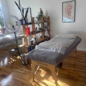 A different view of Frankie’s studio space from another perspective. The two massage tables are visible, but the table on the left side of the photo is slightly cut off. The massage tables are separated by a shelf and room divider with lots of plants, books and other acupuncture supplies on it. Hanging on the wall at the head of the massage table on the Left is a diagram of auricular acupuncture points. The sun is shining into the space from the windows on the left wall. The space is bright and clean.