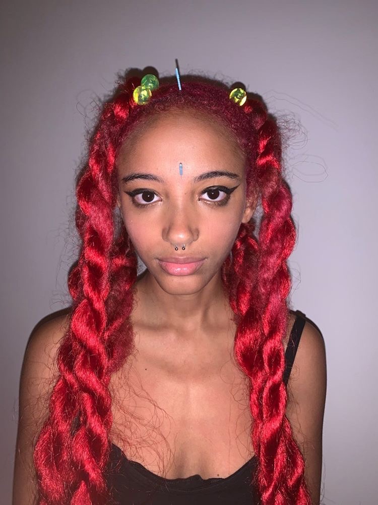 A young black person with long, red twisted hair is looking at the camera. They have an acupuncture needle sticking out from their forehead between their eyes and another one sticking out from the top of their head. They are wearing a black tank top and black winged eyeliner.