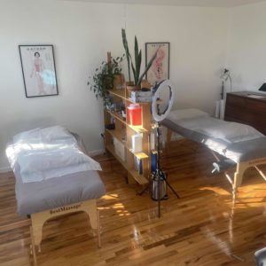 A view of Frankie’s home studio. There are two massage tables set up with gray sheets and white disposable table covers. There are two acupuncture charts hanging on the wall at the head of each table. A room divider and shelf stands between the tables to separate them. The shelf is full of plants, books and other various acupuncture supplies. There is a ring light next to the room divider. The left side of the room is lined with windows and the sun is shining into the space. There is a standing paper lamp and a small acrylic table in the left corner. There is a chest of drawers on the right side of the room.